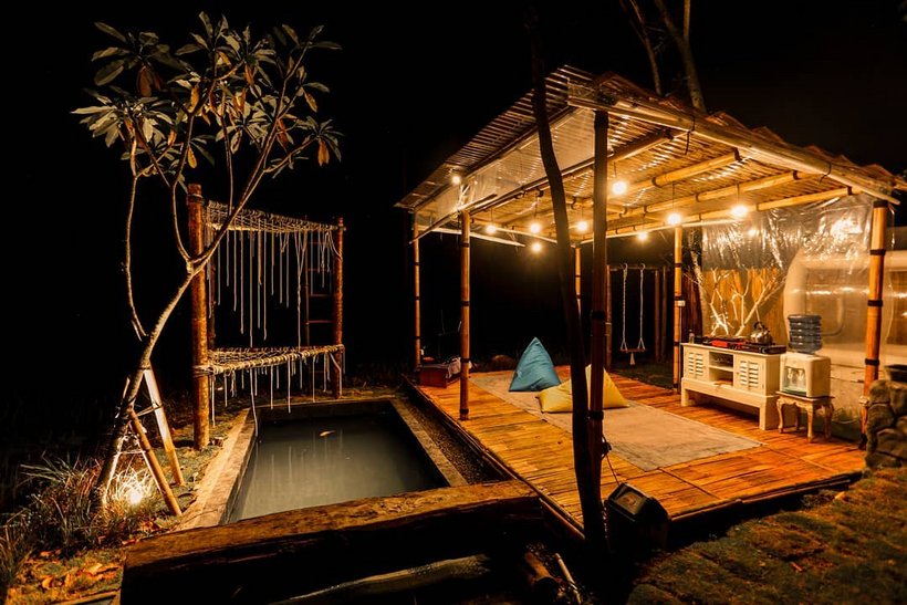 A nighttime view of a wooden deck and thatched-roof structure strung with warm string lights, beside a dark reflective pool