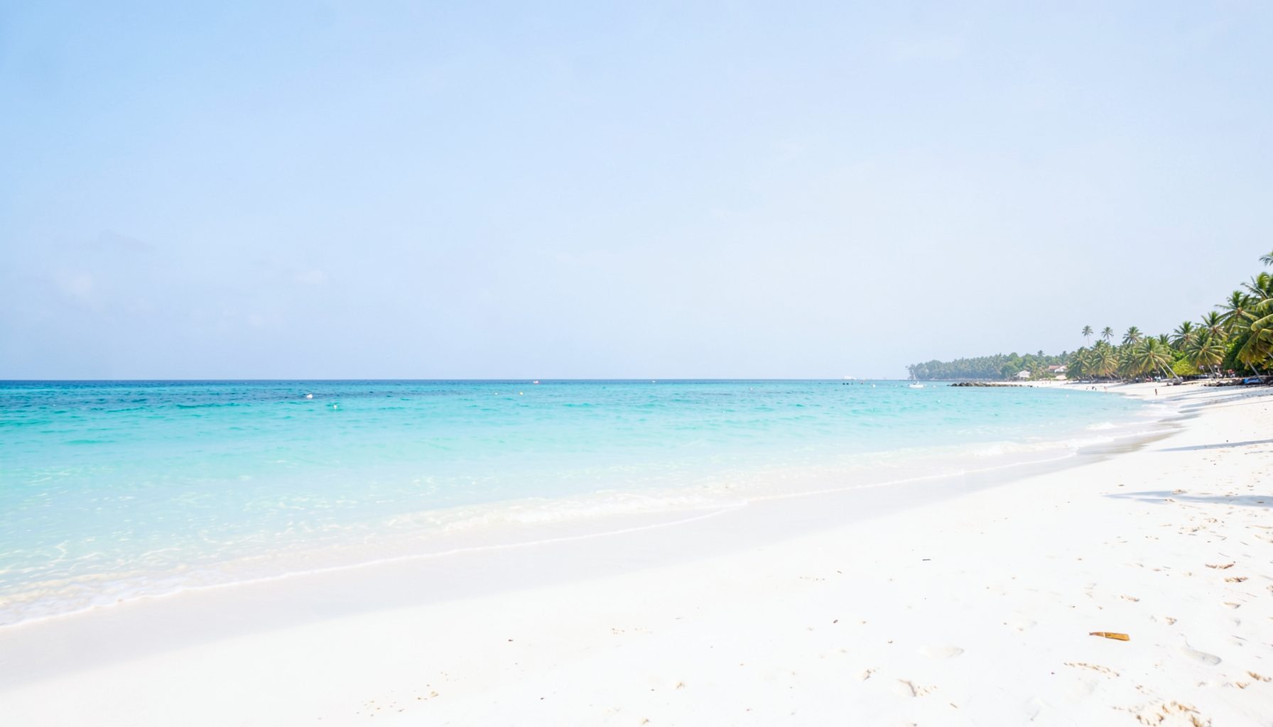 A wide-angle view of a tropical beach with turquoise water and soft white sand under a pale blue sky, calm and inviting