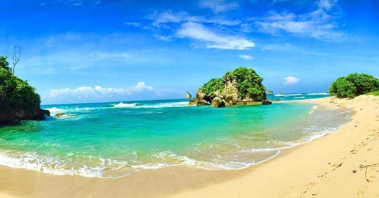 Tropical beach with turquoise water, sandy shore, and a small rocky islet covered in greenery under a blue sky