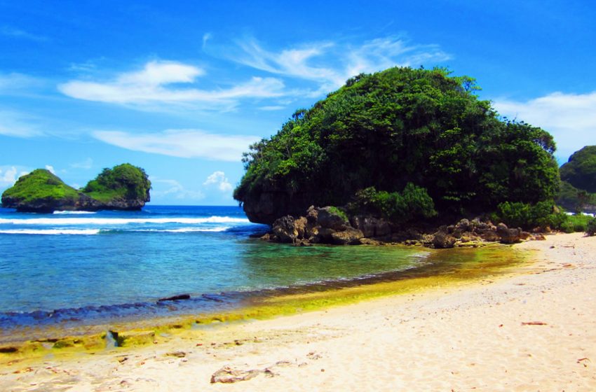A rocky, tree-covered outcrop jutting into turquoise water beside a sandy beach, with smaller islets visible in the back