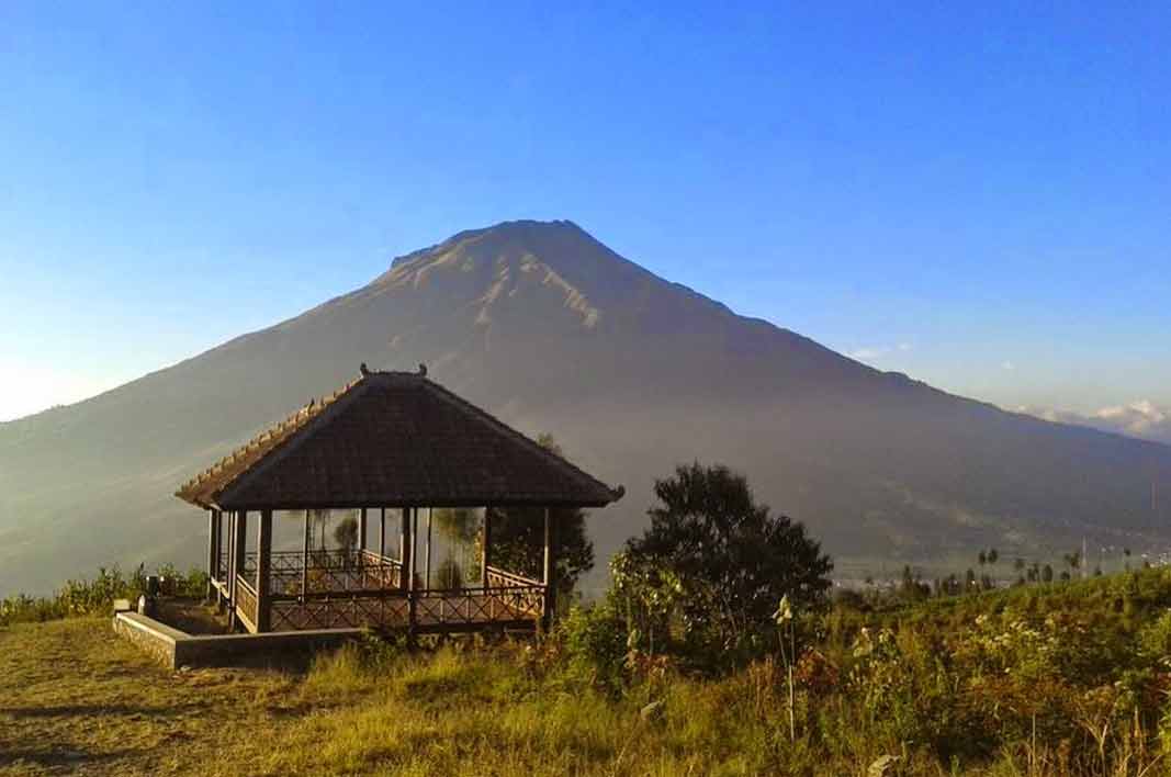 Wooden open-sided pavilion with a tiled roof on a grassy hillside, large volcanic mountain rising under a clear blue sky